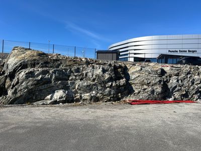 Fotografi av en steinmur eller klippevegg i forgrunnen, med en moderne bygning (Porsche Center Bergen) i bakgrunnen under en klar blå himmel.
