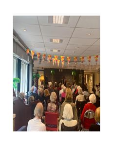 Fotografi av et innendørs arrangement i en stor sal, sannsynligvis i en institusjon eller omsorgsbolig, med publikum og dekorasjoner.