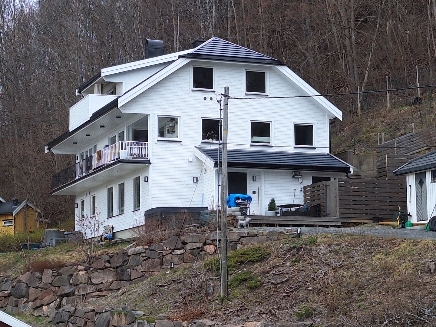 Fotografi av en hvit, moderne villa sett fra siden, med bakgrunn av skog og en steinmur i forgrunnen.