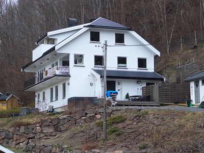 Fotografi av en hvit, moderne villa sett fra siden, med bakgrunn av skog og en steinmur i forgrunnen.