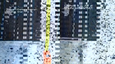 Fotografi av en mørk treplate (sannsynligvis etasjehøyde) på snødekket bakke, med en gul målebånd som strekker seg nedover. Bildet har teksten 'Inngang fasade mot øst' over.