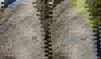 Fotografi av en grusvei eller kjøreflate med en avløpslokk, ved siden av en hekk og en bygning.