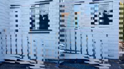 Fotografi av en hvit utvendig fasade med en dør, et vindu og en tilhørende rampe med hvit hekk.