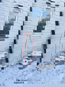 Fotografi av en fasade med hvite panelvegger, et vindu og snø på bakken. Bildet har en håndtegnet markering (rød sirkel) rundt en gul stang ved veggens kant.