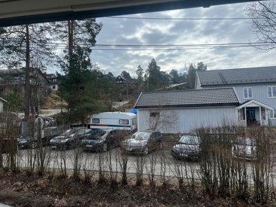 Fotografi av en boligbebyggelse med parkeringsplass, bilvogner og omgivende natur.