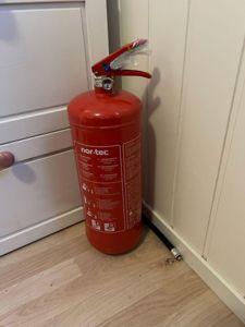 Fotografi av en rød brannslukker (merket 'nor-tec') som står i en hjørne i et rom, med hvite skuffer og panelvegg i bakgrunnen.