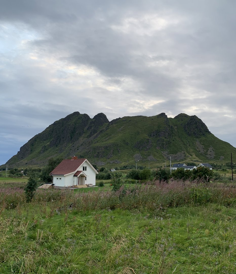 Fotografi av en hvit hytte med rødt tak, plassert i et grønt landskap med en stor fjellformasjon i bakgrunnen.
