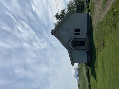 Fotografi av en eksisterende eller ferdigbygd husfasade sett fra siden, med grønt gress og skyet himmel i bakgrunnen.