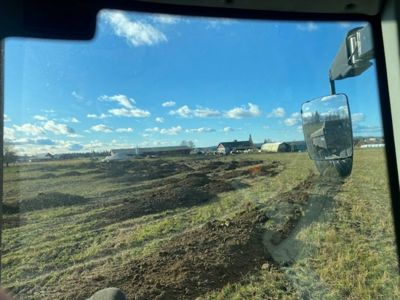 Fotografi tatt fra en kjøretøy (muligens en lastebil eller traktor) som viser et jordbrukslandskap med en grøft eller grav i forgrunnen og bygninger i bakgrunnen.