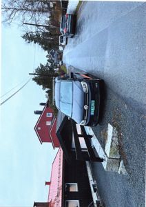 Fotografi av en svart bil parkert foran et hus med rød fasade og et mørkt tak. Bildet viser også en vei, en hage og naboer.