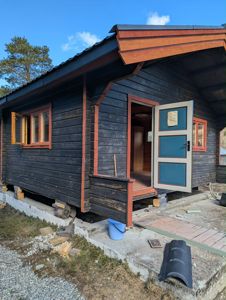 Fotografi av en mørk, trepanelt hytte eller uthus under ombygging eller vedlikehold. Døren står åpen, og det er synlig at bygningen står på betongfundament med mellomrom under.