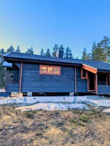 Fotografi av en mørk, trepanelert hytte under oppføring eller renovering, sett fra siden med blå himmel og skog i bakgrunnen.