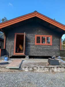 Fotografi av en mørk, trepanelert hytte under pågående utvendig arbeid. Bildet viser fasaden med et åpent døråpning, et vindu, og byggematerialer på forgrunnen.