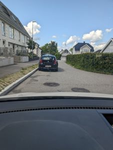 Fotografi tatt fra bil som viser en gate med hus, en parkert bil og en hegg.