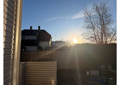Fotografi av en terrasse eller bakgård med en nabohus i bakgrunnen, sett mot solnedgang.