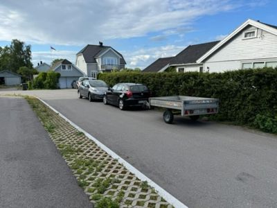 Fotografi av en gate med parkerte biler og boliger i bakgrunnen.