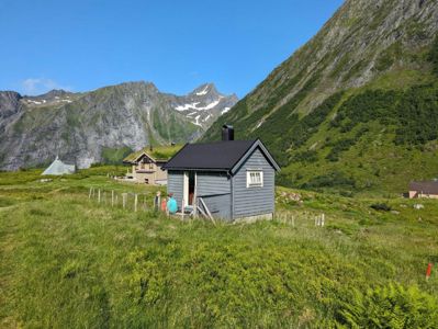Fotografi av en liten hytte i et fjellandskap med to personer foran.
