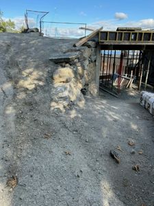 Fotografi av en byggeplass som viser en steinmur, grusvei, og deler av en konstruksjon i gang.