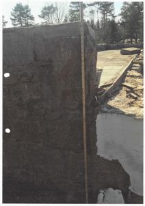 Fotografi av en steinmur under bygging eller restaurering, med målebånd og omgivende terreng.