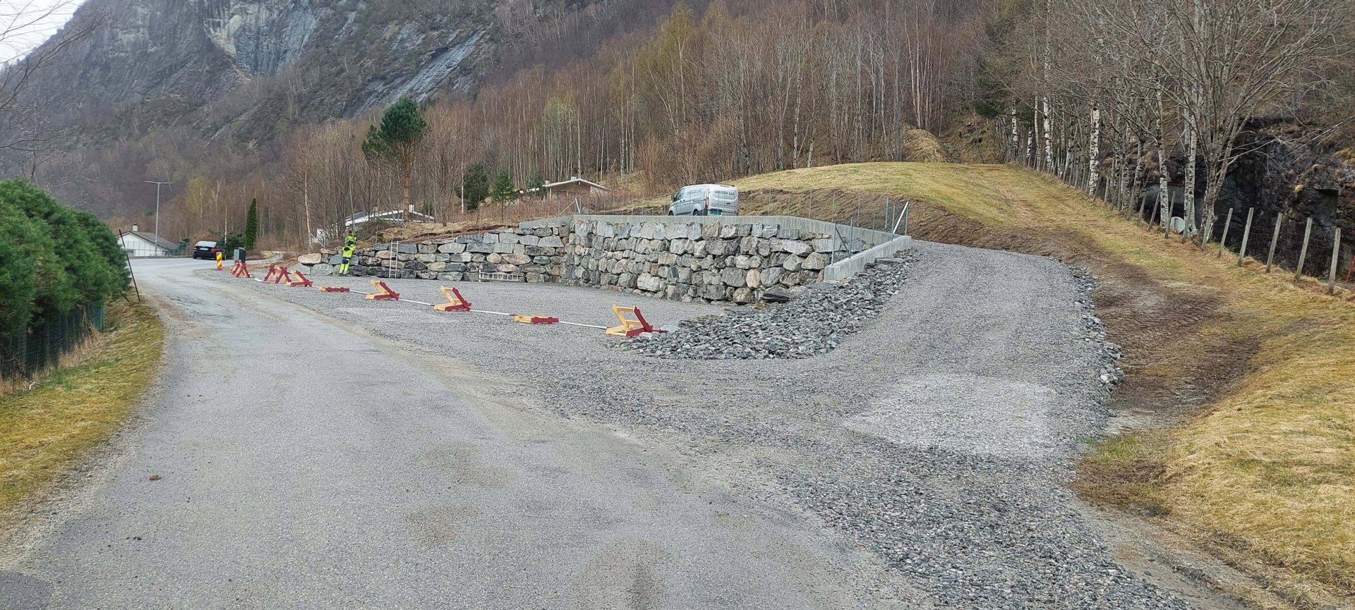 Fotografi av en vei eller tilkomstvei med en steinmur, grus og trafikkregulering i et fjellområde.