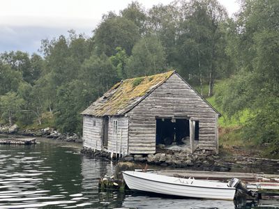 Fotografi av en gammel, forfallelig trehytte på en holme eller kai i vann, med en båt foran og skog i bakgrunnen.