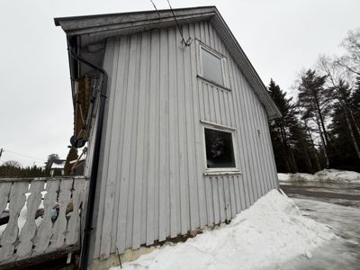 Fotografi av en grå, trepanelt bygning sett fra siden. Bildet viser fasaden, vinduer, takkant, nedløp og en del av en veranda med balustrade. Det er snø på bakken.