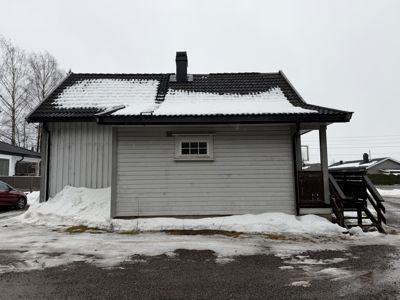 Fotografi av en liten, hvit malte bygning (kanskje en uthus eller garasje) med snø på taket og i omgivelsene.