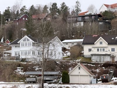 Fotografi av flere hus og garasjer på en skråning, med snø på bakken og trær i forgrunnen.