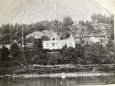 Et gammelt svart-hvitt fotografi som viser en bygning (hus) plassert på et kupert terreng ved vannet, med skog og andre mindre bygninger i bakgrunnen.