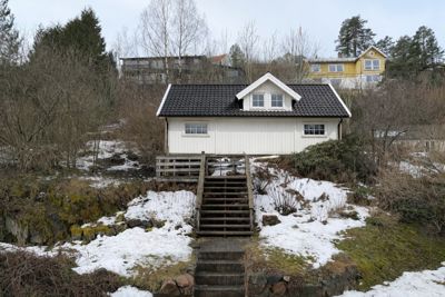 Fotografi av en hvit hytte med mørk takflate, sett fra nede på en skråning med snø og trapper opp til huset.
