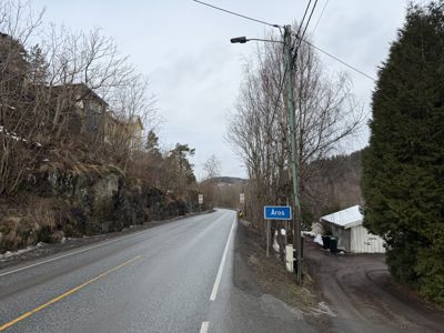 Fotografi av en vei med et skilt som viser navnet 'Åros', samt hus og natur i bakgrunnen.