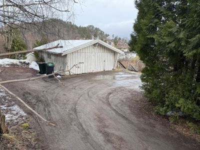 Fotografi av en hvit skur eller garasje sett fra utsiden, med snø på bakken og trær i forgrunnen.