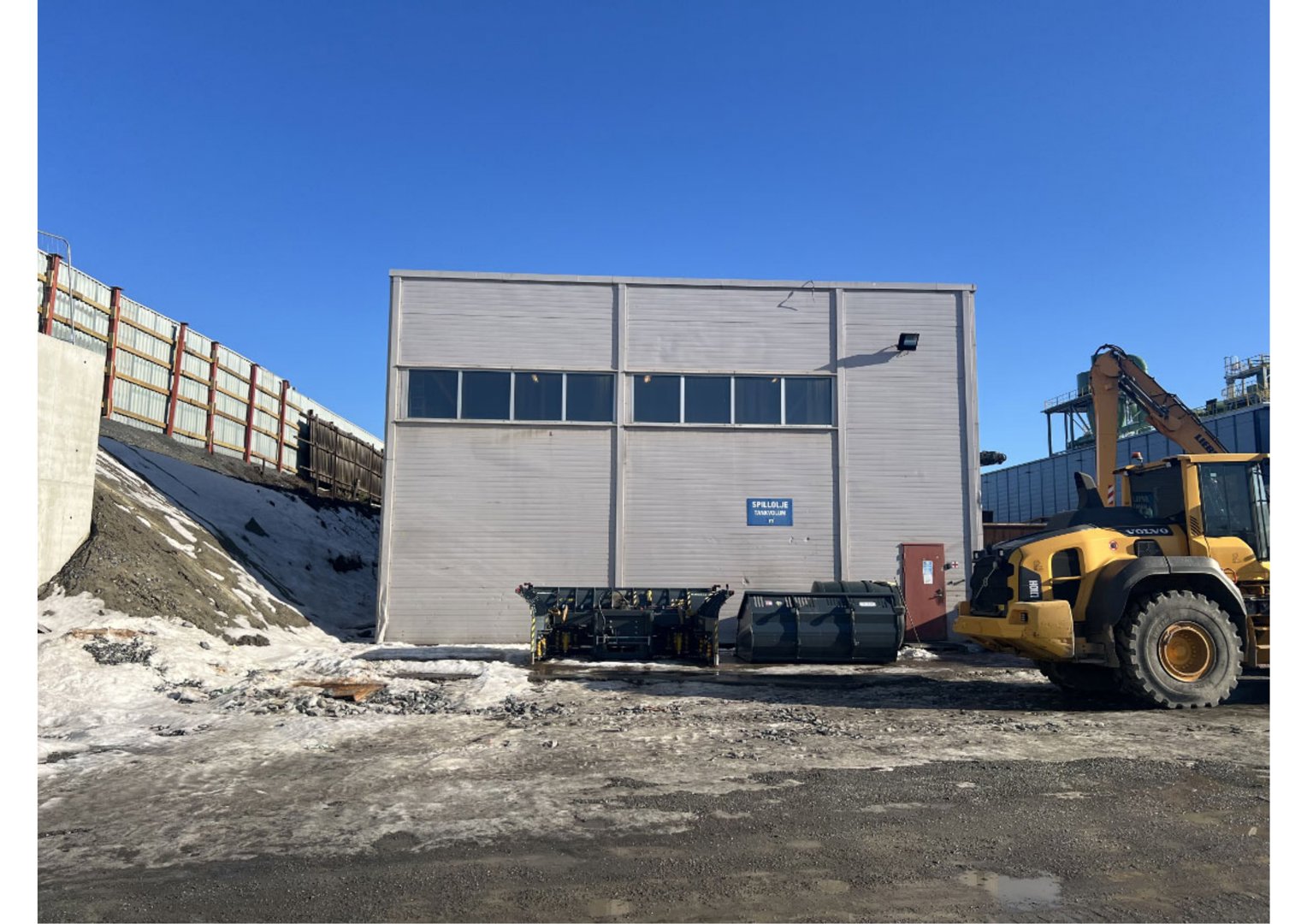 Fotografi av en grå industribygning sett fra fronten, med snø på bakken og en gulelastbil til høyre.
