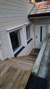 Fotografi av en bygning under oppussing eller nybygging, som viser fasade med trepanel, vinduer, dør og trapper.