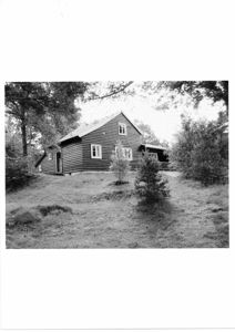 Sort-hvitt fotografi av en liten, trebeslått hytte eller hus sett fra siden, omgitt av trær og grøft.