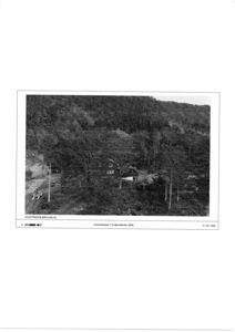 Sort-hvitt fotografi av en eksisterende bebyggelse (hus) sett fra luften, omgitt av skog og terreng.