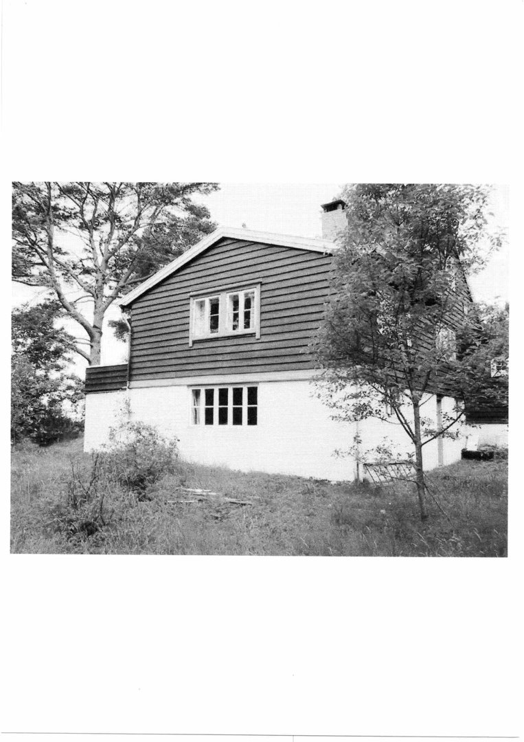 Sort-hvitt fotografi av en toetasjes bygning med skifer- eller trepanel på øvre etasje og hvit fasade på nederste etasje, sett fra siden.