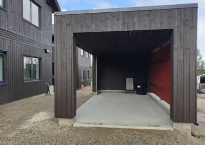 Fotografi av en nybygd, mørk trekonstruksjon (kariport eller garasje) med en betongplate, sett fra utsiden.