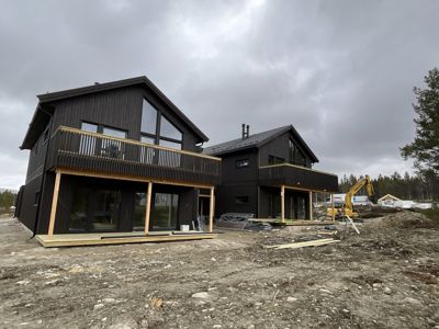 Fotografi av to moderne, mørke trehus under bygging på en tomme med grus og utstyr.