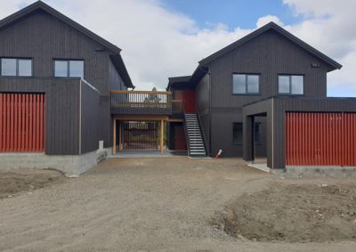 Fotografi av to moderne, mørkebrune hytter med rød detaljering og en betonggrunn. Bildet viser fasaden og den umiddelbare omgivelsene (grusvei) på en byggeplass.