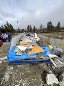 Fotografi av en blå søppelcontainer fylt med byggavfall (treplater, fliser, poser) på en byggeplass. I bakgrunnen ser man en bil med 'NORDBOHUS'-logo og et landskap.