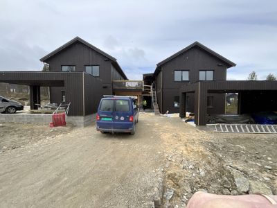Fotografi av en byggeplass med to mørke trehus under oppføring, med bil og byggemateriale i forgrunnen.