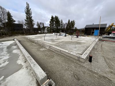 Fotografi av en byggeplass som viser en nylig støpt betongfundamentplate med kantmur, omgitt av grus og skog.
