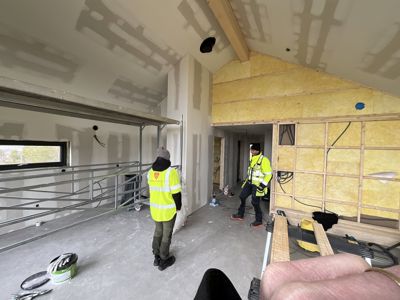 Fotografi av et bygg under oppføring eller renovering, der to personer i sikkerhetsvester er til stede i et rom med ufullførte gipsvegger og gulv.