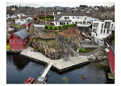 Fotografi av en eksisterende bygning (hvit villa) sett fra luften, med omgivende natur, nabohus og vann.