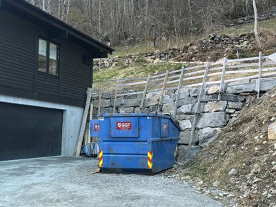 Fotografi av en mørk bygning med garasjeport, en blå søppelcontainer og en steinmur på bakken.