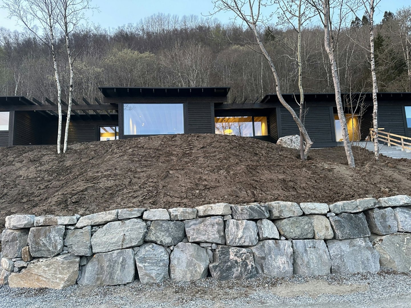 Fotografi av en moderne, mørk bygning sett fra siden, med en steinmur i forgrunnen og en skogkledde bakke i bakgrunnen.