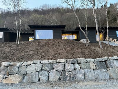 Fotografi av en moderne, mørk bygning sett fra siden, med en steinmur i forgrunnen og en skogkledde bakke i bakgrunnen.