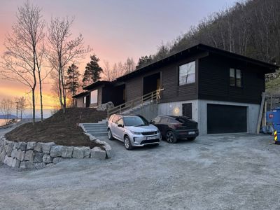 Fotografi av en nybygd eller ferdigstilt hus med mørk fasade, garasje og biloppstillingsplass, tatt mot solnedgang.