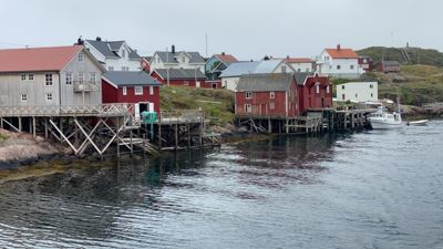 Fotografi av en kystby med flere hus på stoler, en brygge og en båt.